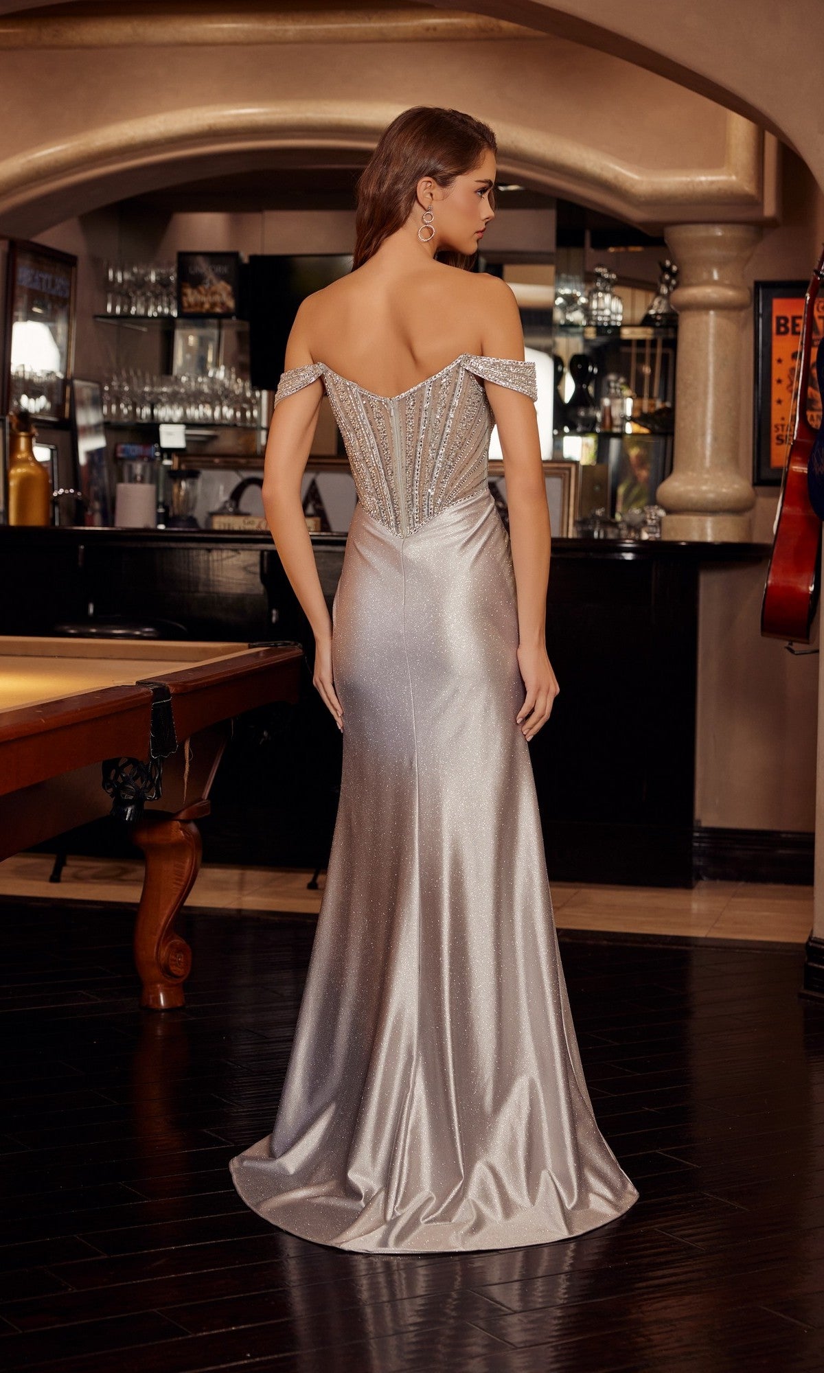A woman wearing the Nox Anabel F1727 long prom dress, featuring a shimmering off-the-shoulder corset bodice, stands with her back to the camera in an elegant bar setting.
